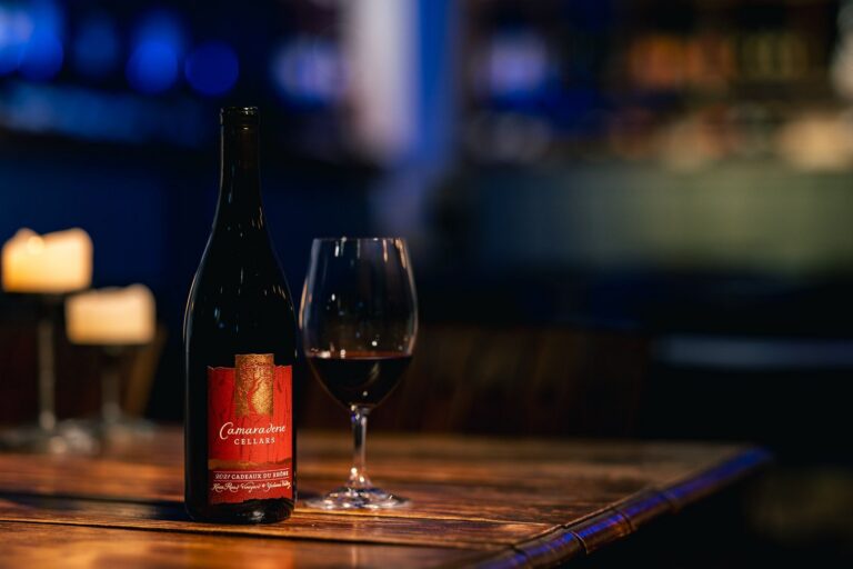Camaraderie Cellars wine bottle and glass on a wooden table with candles and a blurred background.