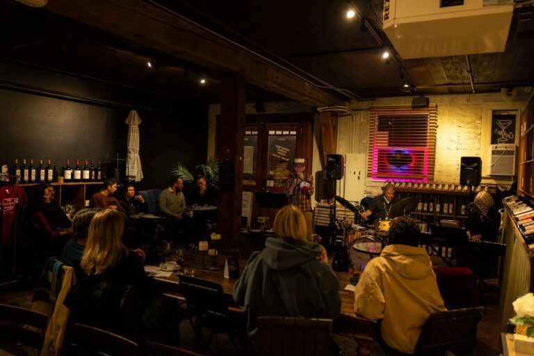 Cozy venue with live band performing to an engaged audience, surrounded by wine bottles and warm lighting.