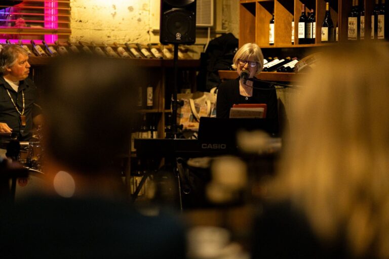 Musician performing on keyboard with a microphone in a cozy, dimly lit venue surrounded by wine bottles.