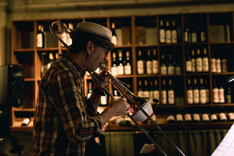 Musician playing a trombone in a cozy venue surrounded by shelves of wine bottles.