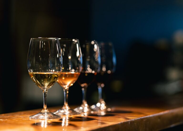 Four wine glasses with different types of wine on a wooden surface in dim lighting.