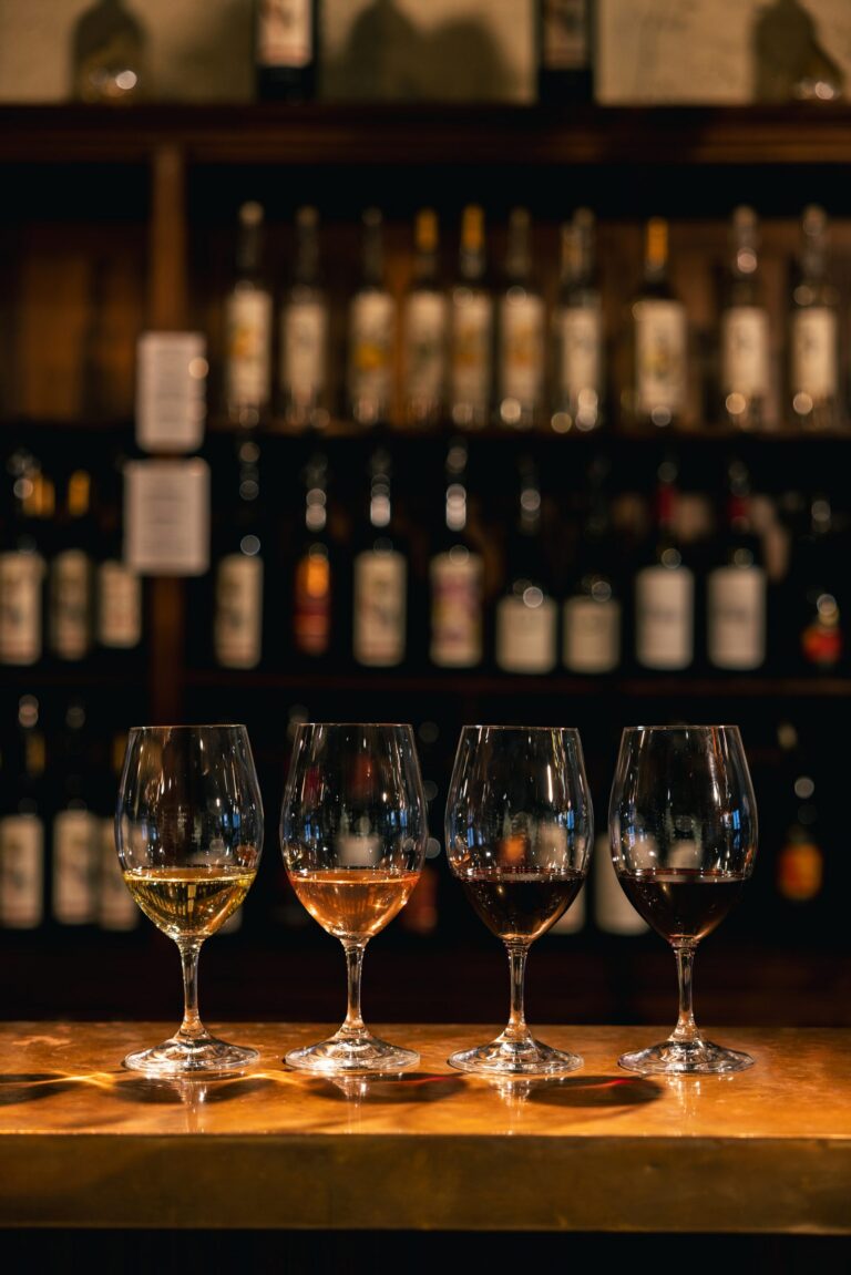 Four wine glasses with different wines on a wooden bar, set against a backdrop of a dimly lit wine shelf.